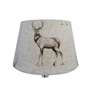 May include: A beige lampshade featuring a brown deer print. The deer is standing on a beige background with a subtle texture.