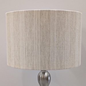 May include: A cylindrical lampshade featuring a light beige, textured fabric with vertical stripes. The stripes alternate between plain bands and raised, braided-style woven patterns. The shade is mounted on a smooth, metallic silver base.