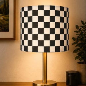 May include: A table lamp featuring a cylindrical lampshade with a black and white chequered pattern. The lamp has a slender, vertical post and a circular base, both finished in a brushed gold metallic tone, emitting a warm, soft glow.