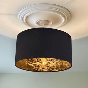 May include: A black drum lampshade with a gold leaf interior hangs from a white ceiling rose. The lampshade's exterior is a solid black, contrasting with the intricate gold pattern inside. This modern lighting fixture adds a touch of elegance.