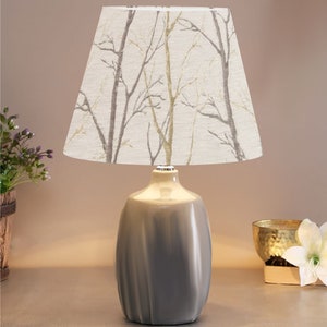 May include: A gray ceramic table lamp with a white lampshade featuring a beige and gray tree branch pattern.