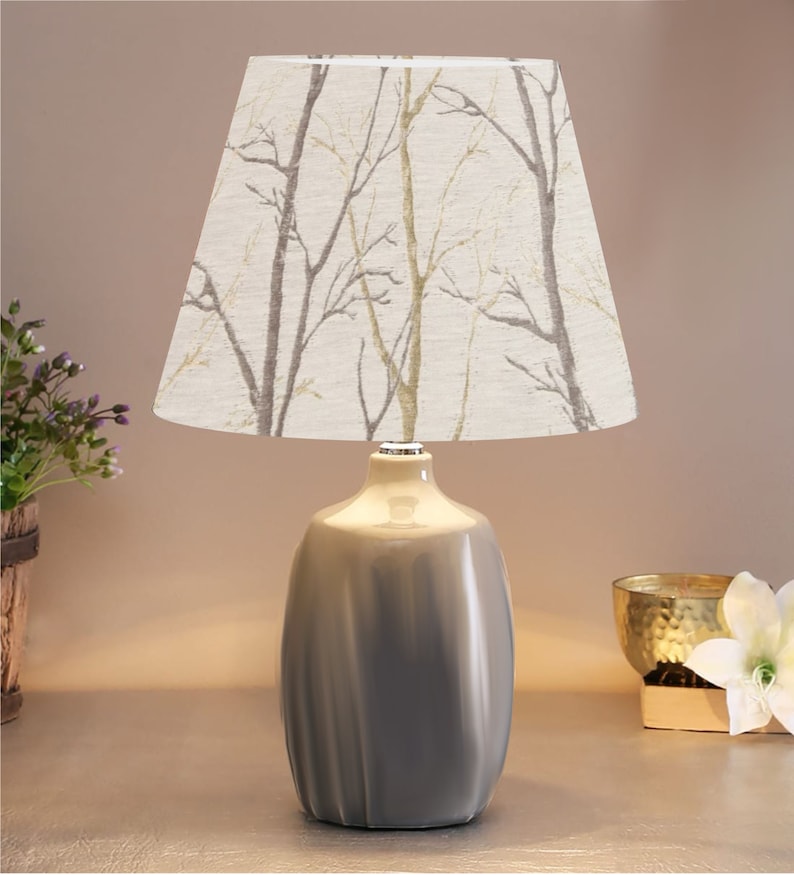 May include: A gray ceramic table lamp with a white lampshade featuring a beige and gray tree branch pattern.