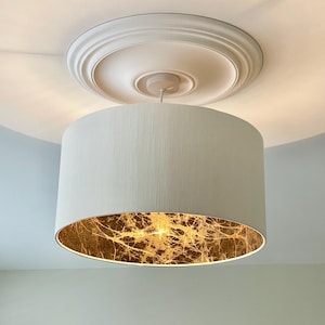 May include: A cream-coloured drum lampshade with a gold leaf interior hangs from a white ceiling medallion. The lampshade's interior reflects a warm, golden light. The ceiling is white, and the room has a neutral colour palette.