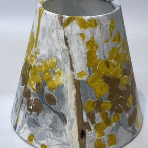 May include: A small, tapered lampshade with a fabric design featuring yellow, brown, and grey floral and tree branch patterns on a white background. The lampshade is designed for a small lamp base.