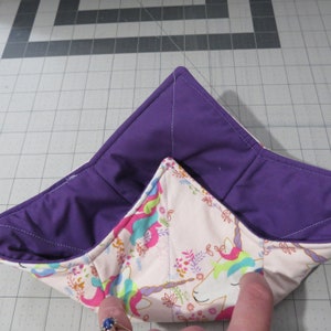 May include: A purple and pink fabric bowl with a unicorn print. The bowl is folded in half, showing the purple lining.