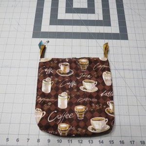 May include: A brown and white quilted coffee-themed pot holder with a repeating pattern of coffee cups, coffee beans, and coffee-related words. The pot holder has a loop for hanging and is secured with a safety pin.