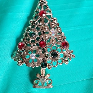 May include: A silver Christmas tree brooch with red and green rhinestones. The tree is decorated with a star at the top and has a detailed filigree design.