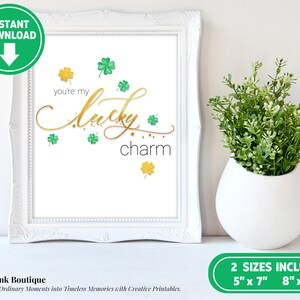 May include: Printable art print with gold lettering that says "you're my lucky charm" with green shamrocks scattered around the text. The print is framed in a white frame.
