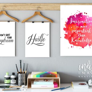 May include: Three motivational posters with white backgrounds and black text. The posters are hanging on a wall with a wooden shelf below. The posters read "Don't Quit Your Daydream", "Hustle", and "Imagination is more important than knowledge".