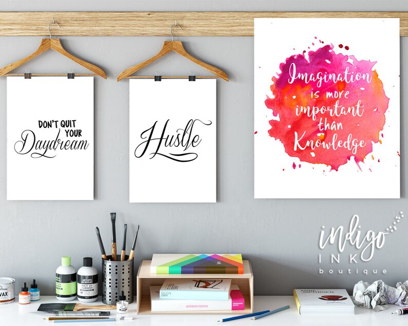 May include: Three motivational posters with white backgrounds and black text. The posters are hanging on a wall with a wooden shelf below. The posters read "Don't Quit Your Daydream", "Hustle", and "Imagination is more important than knowledge".