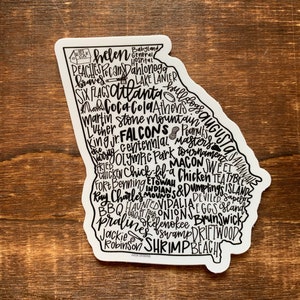 May include: A white sticker with a black outline of the state of Georgia. The sticker is filled with text, including the names of cities, landmarks, and attractions in Georgia. Some of the text includes: Atlanta, Augusta, Savannah, Helen, Dahlonega, Stone Mountain, and the Georgia Bulldogs.
