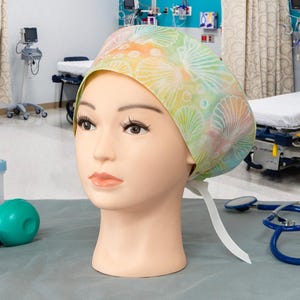 May include: A scrub cap featuring a pastel-colored pattern of seashells and bubbles on a light background. The cap is worn on a mannequin head and includes white fabric ties at the back to secure the fit, providing a functional head covering.