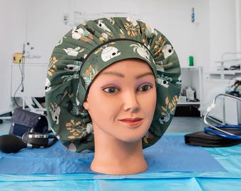 May include: A green scrub cap with a skull and floral pattern is displayed on a mannequin head. The cap has a black elastic band and is set against a medical setting with medical equipment in the background.