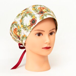 May include: A white scrub cap featuring a fall-themed pattern with pumpkins, sunflowers, and foliage. The cap is tied with a burgundy ribbon. The cap is displayed on a mannequin head.