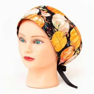 May include: A patterned scrub cap featuring a black background with orange and white pumpkins, berries, and a small bird. The cap is tied with a black ribbon. The cap is displayed on a mannequin head.