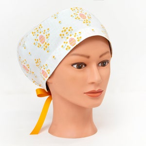 May include: White surgical scrub cap with a floral pattern featuring yellow and pink flowers. The cap has a yellow tie.