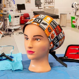 May include: A mannequin head wearing a Halloween-themed surgical scrub cap. The cap features a patchwork design with orange, black, white, and gray squares with Halloween icons. A stethoscope and medical instruments are in the foreground.