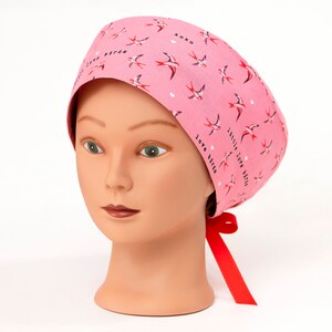Puede incluir: Un gorro de quirófano rosa con un lazo rojo. El gorro tiene un patrón de pájaros blancos con detalles rojos y el texto "Love Birds" y "Little Love Birds" repetido en la tela.