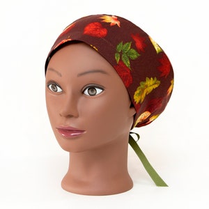 May include: A brown surgical scrub cap with a repeating pattern of autumn leaves in red, yellow, and green. The cap is tied at the back with a green ribbon. The cap is displayed on a mannequin head.