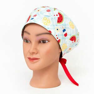 May include: A white scrub cap with a repeating pattern of red watermelon slices, yellow lemonade, and red, white, and blue popsicles. The cap has a red tie in the back and is displayed on a mannequin head.
