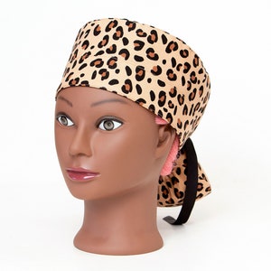 May include: A brown and black leopard print surgical scrub hat with a black tie. The hat is on a mannequin head.