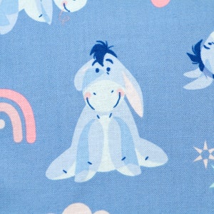 May include: Light blue fabric featuring a repeating pattern of Eeyore from Winnie the Pooh. The design includes pink rainbows, stars, and clouds. The fabric is suitable for various crafting projects.