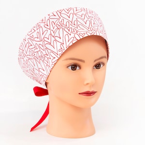 May include: A white scrub cap with a repeating pattern of red hearts. The cap is tied in the back with a red ribbon. The cap is displayed on a mannequin head.