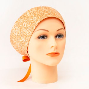 May include: An orange and white patterned scrub cap with a matching orange ribbon tie. The cap is displayed on a mannequin head against a white background. The cap features a paisley-like design.