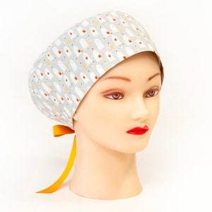 May include: A light gray scrub cap featuring a repeating pattern of white ghost figures with orange and yellow accents. The cap is tied with a bright yellow ribbon. The cap is displayed on a mannequin head with red lipstick.
