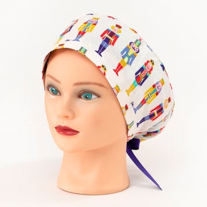 May include: White scrub cap with a colorful pattern of toy soldiers. The cap is tied with a purple ribbon. The toy soldiers are in red, blue, yellow, and green. The cap is on a mannequin head.