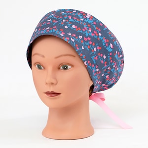 May include: A dark blue surgical scrub cap with a floral pattern in pink and blue. The cap is tied in the back with a light pink ribbon. The cap is displayed on a mannequin head.