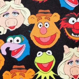 May include: A black fabric featuring a repeating pattern of Muppet character faces including Kermit the Frog, Miss Piggy, Fozzie Bear, Gonzo, and Animal. Each character is rendered in bright, distinct colors with detailed facial expressions.