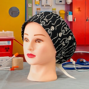 May include: A black skull print surgical scrub cap on a mannequin head. The cap features a repeating pattern of white skeletons. Medical supplies, including syringes, alcohol pads, and a stethoscope, are in the background.