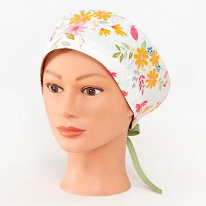 May include: White floral patterned scrub cap featuring yellow, pink, and blue flowers with green leaves. The cap has a light green ribbon tie at the back. The cap is on a mannequin head.