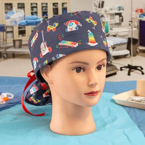 May include: A blue scrub hat with a colorful pattern of toys, including a toy phone, a toy xylophone, a toy truck, and a stacking toy. The hat is on a mannequin head.