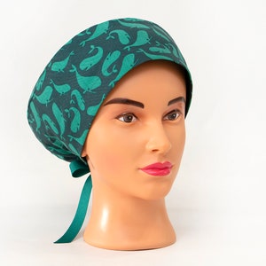 May include: A teal and dark teal patterned scrub cap featuring a whale design. The cap is tied at the back with a teal ribbon. The cap is displayed on a mannequin head against a white background.