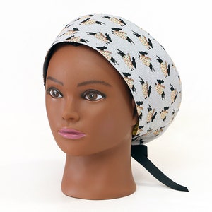 May include: A light blue surgical scrub cap with a repeating pattern of black and tan llamas. The cap is tied in the back with a black ribbon. The cap is on a mannequin head.