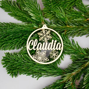 Puede incluir: Un adorno navideño de madera con el nombre "Claudia" recortado en el centro. El adorno está decorado con copos de nieve y tiene un acabado dorado.