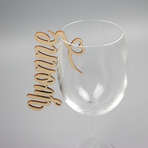 May include: A wooden wine glass charm with the word "Grandma" written in cursive. The charm is attached to a wine glass.