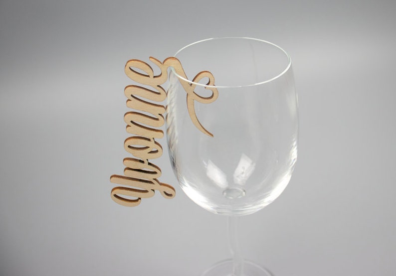 May include: A wooden wine glass charm with the word "Grandma" written in cursive. The charm is attached to a wine glass.