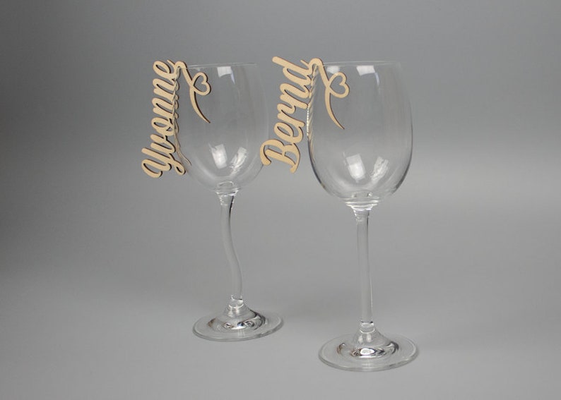 May include: Two wine glass charms with the names "Yvonne" and "Bernd" written in cursive on them. Each charm has a small heart at the end of the name.