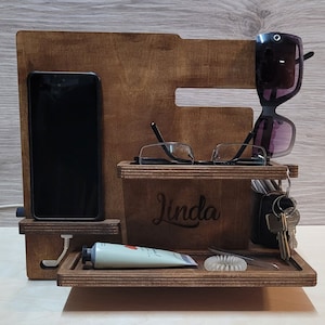 Może przedstawiać: Drewniany organizer z podstawką na telefon, przegrodą na okulary, półką na klucze i małą tubką kremu. Organizer jest spersonalizowany imieniem "Linda".