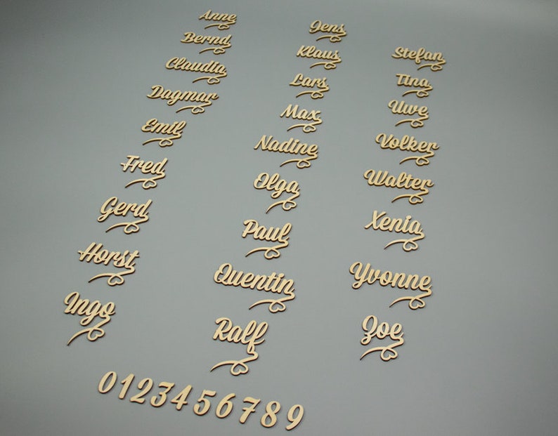 May include: Wooden name place cards with cursive lettering in gold. The names include Anne, Bernd, Claudia, Dagmar, Emil, Fred, Gerd, Horst, Ingo, Jens, Klaus, Lars, Max, Nadine, Olga, Paul, Quentin, Ralf, Stefan, Tina, Uwe, Volker, Walter, Xenia, Yvonne, and Joe. The numbers 0 through 9 are also included.