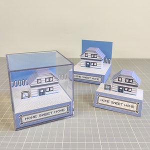 Może przedstawiać: Trzy miniaturowe domki z papieru z napisem "Home Sweet Home" na podstawie. Domki są białe z niebieskimi ozdobami i mają pikselowany wzór. Jeden domek znajduje się w przezroczystym plastikowym sześcianie.