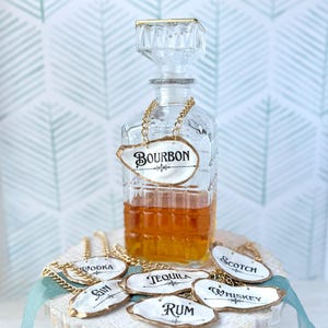 May include: A clear glass bourbon decanter with a crystal stopper and a gold chain label that reads "Bourbon." Additional labels for other liquors are displayed on a white, distressed wooden stand. The labels have gold trim and black lettering.