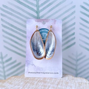 May include: A pair of handcrafted earrings made from mussel shells. The earrings have a gold-colored trim and a pearlescent interior with blue and gray hues. The earrings are displayed on a white card with the text "thesouthernoysterco.com".