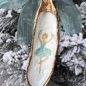 May include: A decorative, elongated ornament with a gold-painted rim and a white interior. The ornament features a watercolor illustration of a ballerina in a blue tutu. A light blue ribbon is attached to the top, and the ornament is displayed on a snowy evergreen branch.