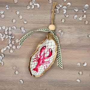 May include: A gold-painted oyster shell ornament with a red lobster illustration inside. The ornament has a green and white ribbon and a wooden bead for hanging.