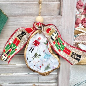 May include: A white oyster shell ornament with a gold rim, decorated with a nutcracker, a ballerina, and other festive details. The ornament is tied with a red and green ribbon with a gold trim and a nutcracker design.