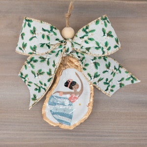 May include: A gold oyster shell ornament featuring a painted image of a woman in a blue and white dress. The ornament is decorated with a white and green bow with gold trim.